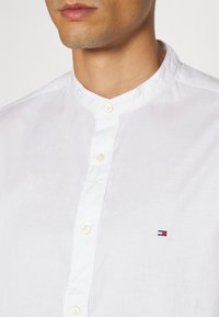 Camicia bianca in cotone con collo a band, dotata di chiusura a bottoni e un piccolo logo ricamato in blu, rosso e bianco sul petto.