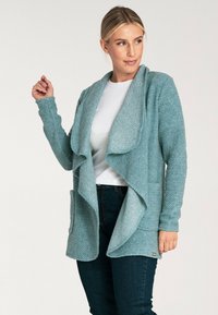 Jasnoniebieski kardigan zapinany z przodu, wykonany z teksturowanej tkaniny, z długimi rękawami i dwiema przednimi kieszeniami. Miękka konstrukcja bez zapięć.