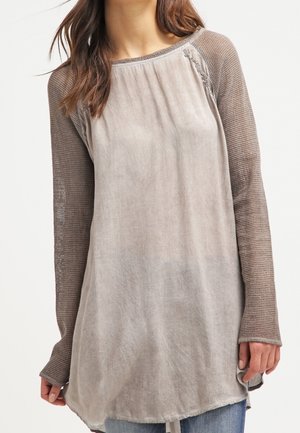 Vrouw draagt een losse beige top met getextureerde gebreide lange mouwen en lichtblauwe jeans, staande tegen een effen witte achtergrond.