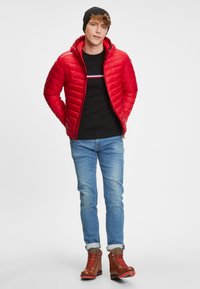 Rød pufferjakke med hætte, quiltet struktur og lynlås, parret med en sort sweater og blå jeans. Brune snørebåndsstøvler fuldender looket.