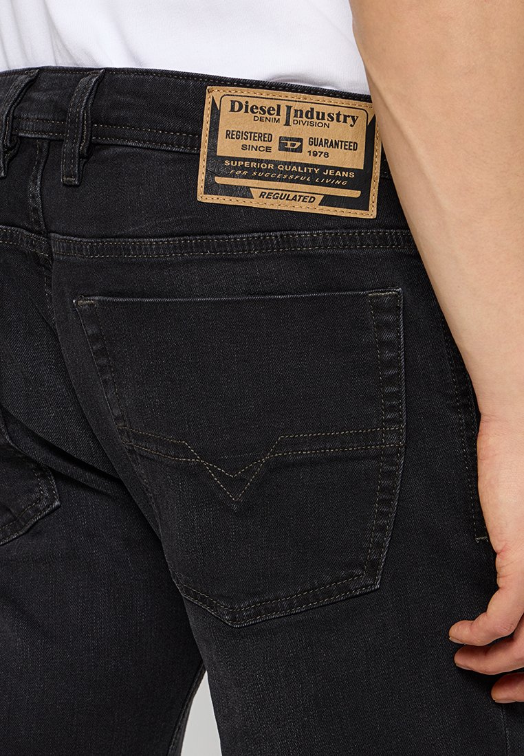 Schwarze Denim-Jeans mit einem braunen Lederetikett mit Text, sichtbaren Nähten und einer Tasche mit einem V-förmigen Design auf der Rückseite.
