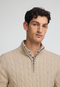 Beige stickad tröja med dragkedja i kragen, med kabelstickad textur och en liten logotyp på bröstet. Lagd över en rutmönstrad skjorta.