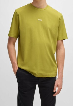 Homme portant un t-shirt uni vert olive à manches courtes avec le mot BOSS en petites lettres blanches sur la poitrine et un pantalon noir.