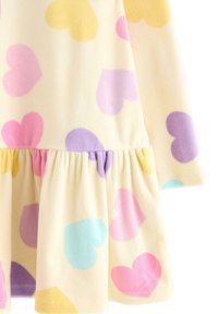 Langærmet kjole i blød, lys gul stof med store, farverige hjerte mønstre i pink, lilla, blå og gul; har en flæse nederste kant.