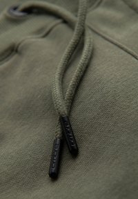 Nærbillede af grønt hættetrøjestof med flettede snore, der ender i sorte snørelåse prydet med "Ombre".