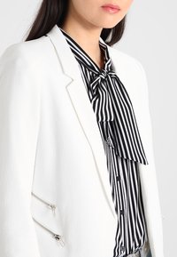 Blazer blanc avec fermetures éclair argentées, porté par-dessus un chemisier rayé noir et blanc noué au cou, avec des boutons et une texture lisse.