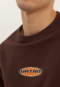 Nærbilde av en person som har på seg en brun t-skjorte med en oransje og svart oval logo med teksten "URTRA SPORTS" på brystet.