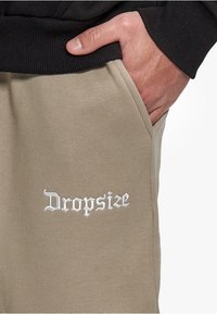 Beige jogger pants with a smooth texture, featuring an embroidered white "Dropsize" logo on the front pocket area.