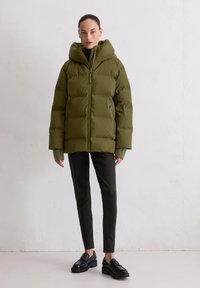 Olijfgroene pufferjack met een capuchon, voorzien van een voorrits, twee zijzakken en ribgebreide manchetten, gecombineerd met zwarte slim-fit broeken en zwarte schoenen.