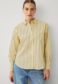 Chemise à manches longues jaune rayée de fines rayures bleu marine, fermeture par boutons et col classique. Tissu doux avec coupe décontractée.