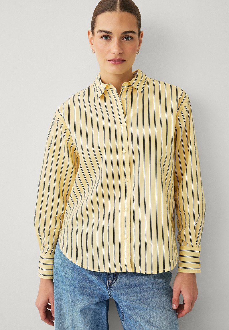 Chemise à manches longues jaune rayée de fines rayures bleu marine, fermeture par boutons et col classique. Tissu doux avec coupe décontractée.