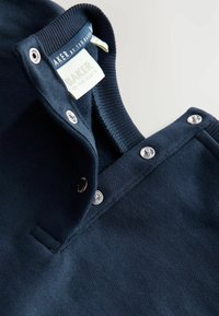 Jūras zilās adītas polo kakla krekls ar sudraba spiedpogu pogām. Iezīmēts ar ribotu apkakli un birku "AKER by Ted Baker." Mīksta tekstūra.
