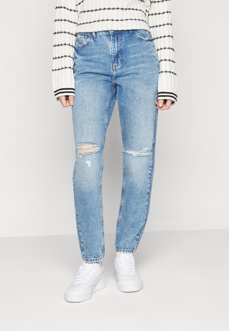Ljust blå högmidjade jeans med slitna lappar, med en lös passform och avsmalnande ben. Kombinerat med vita sneakers och en randig tröja.
