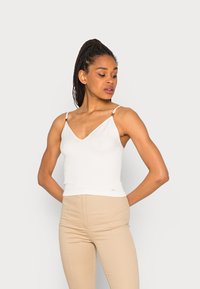 Camisola branca canelada com decote em V e alças ajustáveis, apresentando detalhes em metal dourado. Combinada com calças bejes ajustadas.