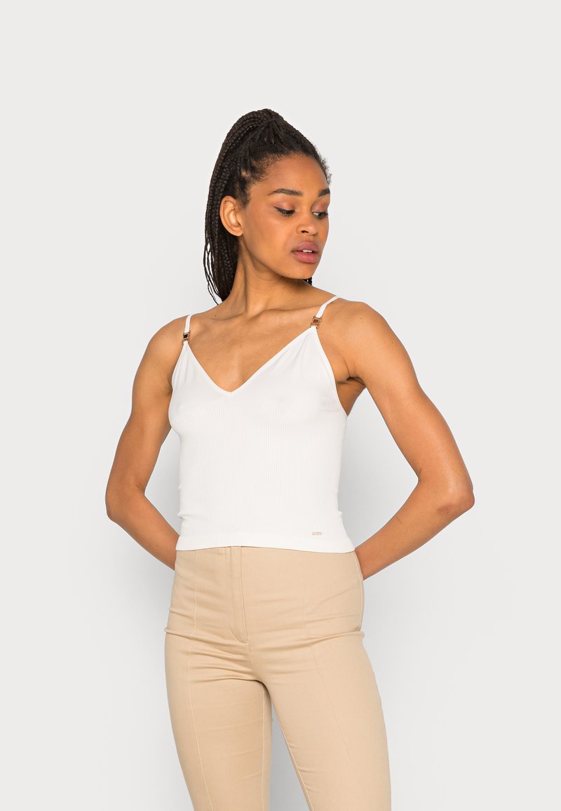 Camisola branca canelada com decote em V e alças ajustáveis, apresentando detalhes em metal dourado. Combinada com calças bejes ajustadas.