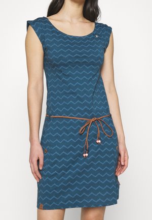 Nainen, jolla on yllään sininen hihaton mekko, jossa on kalanruotokuvio, ruskea punottu vyö solmittu vyötärölle ja siinä vaaleanpunaisia helmikoristeita.