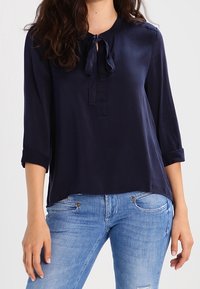 Kvinna klädd i en marinblå blus med 3/4-ärmar och knytkrage, matchad med ljusblå jeans.