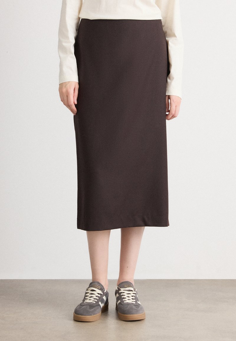 Jupe de maternité en tissu marron foncé, longueur genoux avec un design droit et épuré. Associée à des baskets grises avec une surface texturée.