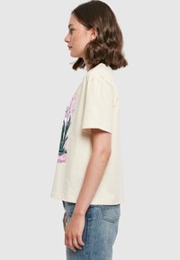 Kremfarget t-skjorte med korte ermer og blomstermotiv i grønn og rosa. Myk bomullsmateriale og løs passform, kombinert med lyseblå jeans.