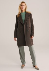 Cappotto marrone doppiopetto con rever a scollatura, due tasche anteriori, indossato sopra un maglione verde testurizzato e pantaloni verdi chiari su misura.
