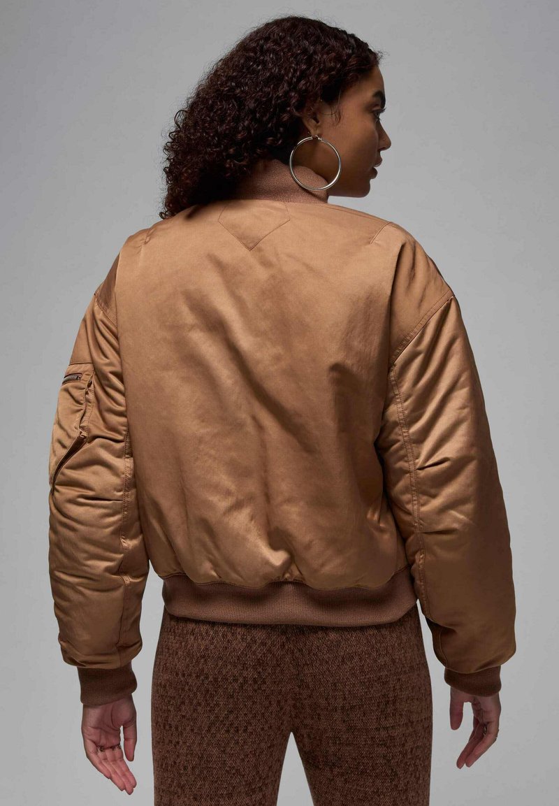 Veste bombardier marron avec une texture lisse et brillante, des poignets côtelés et une poche utilitaire sur la manche ; portée sur un pantalon marron à motifs.