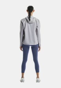 Hellgraue Kapuzenjacke mit elastischen Manschetten, kombiniert mit marineblauen Leggings. Die Jacke hat einen schlanken Schnitt und eine glatte Textur.