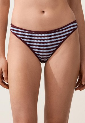 Žena oblečená v rebrovaných bikinových nohavičkách s horizontálnymi svetlomodrými a tmavohnedými pruhmi, stojaca s uvoľnenými rukami po stranách.