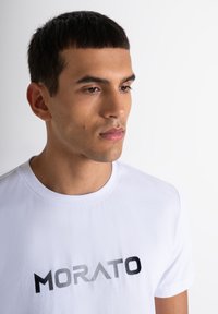 Camiseta de algodón blanca con un logo en degradado negro "MORATO" en el pecho. Mangas cortas y cuello redondo. Textura suave.