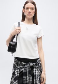 Νεαρή γυναίκα με ίσια μαλλιά, φορώντας λευκό μπλουζάκι και ασπρόμαυρο πουκάμισο καρό δεμένο γύρω από τη μέση, κρατώντας μαύρη τσάντα στον ώμο.