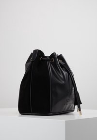 Musta olkalaukku, jossa yhdistyvät sileä nahka ja mokkanahka. Laukussa on kullanväriset metalliosat, kiristysnyörikiinnitys ja tyylikäs kohokuvioitu logodetalji.