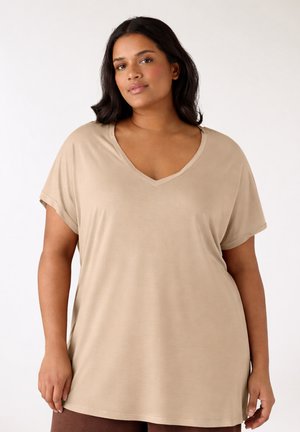 Donna che indossa una maglietta beige con scollo a V e maniche corte, larga, in piedi davanti a uno sfondo chiaro e uniforme.