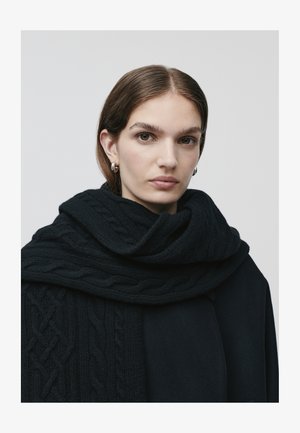 Écharpe en tricot noir avec un motif en relief, enroulée autour du cou, assortie à un manteau noir. Texture lisse et design superposé visibles.