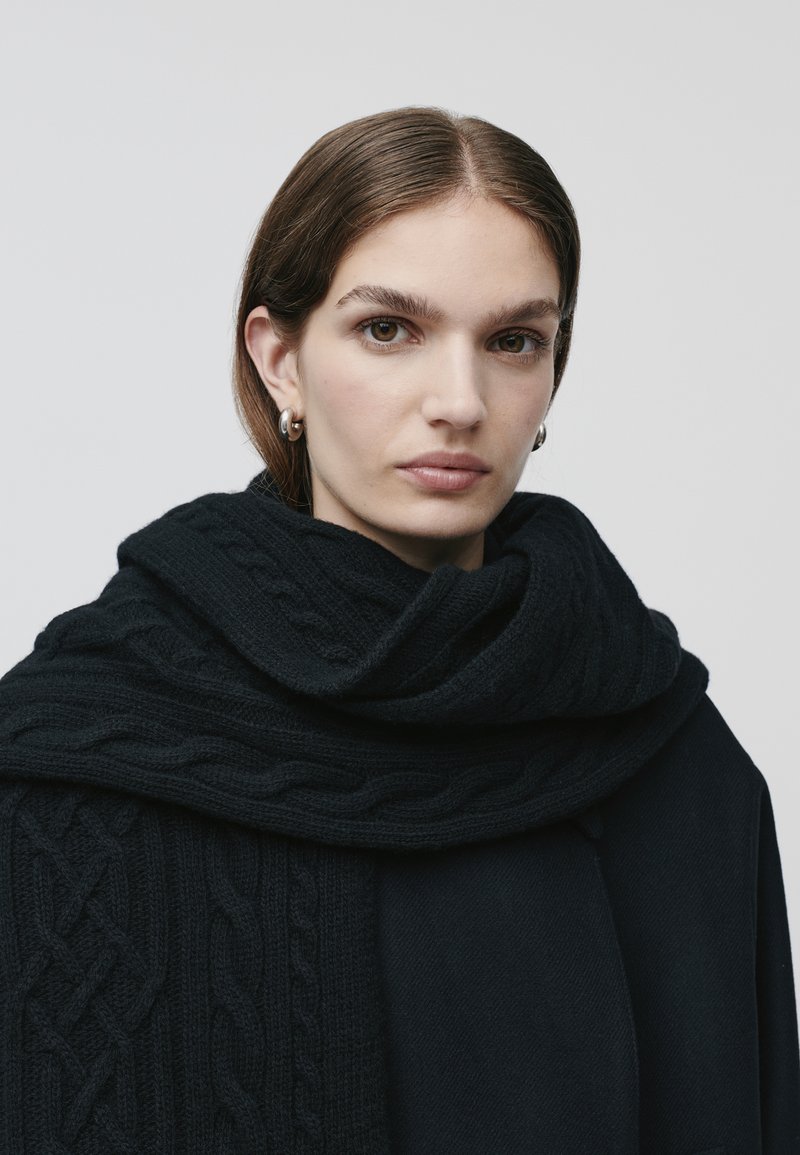 Sciarpa nera lavorata a maglia con motivo a trecce, avvolta attorno al collo, abbinata a un cappotto nero. Texture liscia e design stratificato visibili.