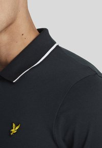 Mørkeblå polo-skjorte med teksturert stoff, hvit kantet krage, og en liten gul ørn-logo på venstre side.