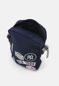 Zaino in tessuto blu navy con chiusura a zip, caratterizzato da vari patch e loghi, tra cui "NEW YORK" e un emblema di baseball.