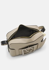 Beige Ledertasche mit Reißverschluss, schwarzen Lederakzenten und einer silbernen Logo-Plakette. Das Innere ist schwarz mit kontrastierenden Nähten.