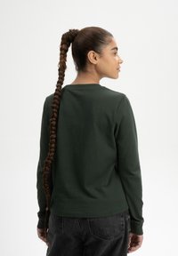 Donkergroen langslavend shirt van zachte stof, met een ronde halslijn en een comfortabele pasvorm. De achterkant toont gevlochten haar en een casual stijl.