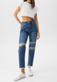Jeans de mezclilla azul de tiro alto con rodilleras desgastadas, con dobladillos deshilachados, combinados con una camiseta corta blanca y zapatillas blancas.