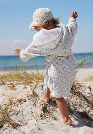 Taaperolla on päällään huppullinen kukkakuosinen kylpytakki ja yhteensopiva hattu, ja hän kävelee paljain jaloin hiekkaisella dyynillä meren rannalla kirkkaan taivaan alla.