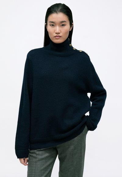 Navy coltrui in een zachte brei, met gouden knopen op één schouder, gecombineerd met geruite grijze broek. Casual ontwerp en een ontspannen pasvorm.