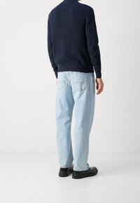 Mörkblå texturerad tröja med ribbade ärmmuddar och nederkant, i kombination med ljusblå avslappnade jeans och svarta skor. Synliga detaljer på baksidan av tröjan.