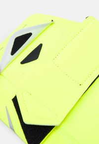 Sangle réfléchissante jaune fluo avec des accents noirs et des surfaces texturées. Dotée d'une fermeture velcro sécurisée et de bords lisses surpiqués.