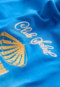 Tissu bleu avec une coquille Saint-Jacques jaune brodée et un texte en cursive blanche indiquant "Club Odiseo".