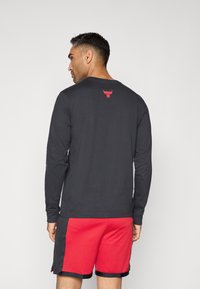 Langärmliges schwarzes Shirt mit rotem Bullenlogo auf dem Rücken, kombiniert mit roten Shorts, die ein strukturiertes Muster und schwarze Akzente aufweisen.
