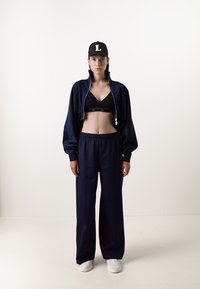 Fato de treino azul marinho com uma jaqueta cropped e calças largas, combinado com um top desportivo preto e sapatilhas brancas. Apresenta um boné com logo.