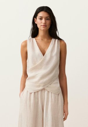 Femme aux longs cheveux bruns portant un haut portefeuille beige sans manches et un pantalon large assorti, sur un fond clair uni.