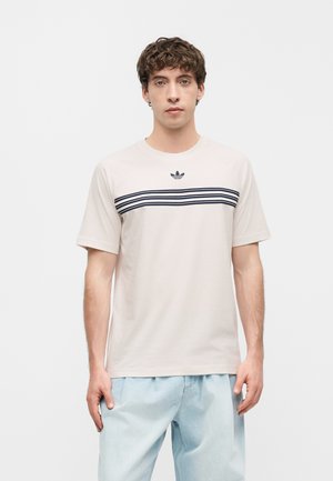Młody mężczyzna w beżowej koszulce Adidas z czarnymi poziomymi paskami i logo, w połączeniu z jasnoniebieskimi jeansami, stoi na jednolitym tle.