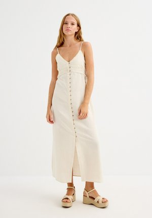 Mujer con vestido maxi sin mangas color crema con botones en la parte delantera, tirantes finos y sandalias de cuña beige sobre fondo blanco liso.