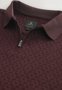 Burgundy polo shirt with a zip collar, featuring a textured geometric pattern and a label showing size medium, brand Next, and regular fit.