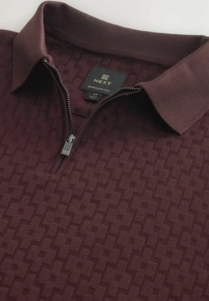 Polo burdeos con cuello de cremallera, que presenta un patrón geométrico texturizado y una etiqueta que indica tamaño mediano, marca Next y corte regular.